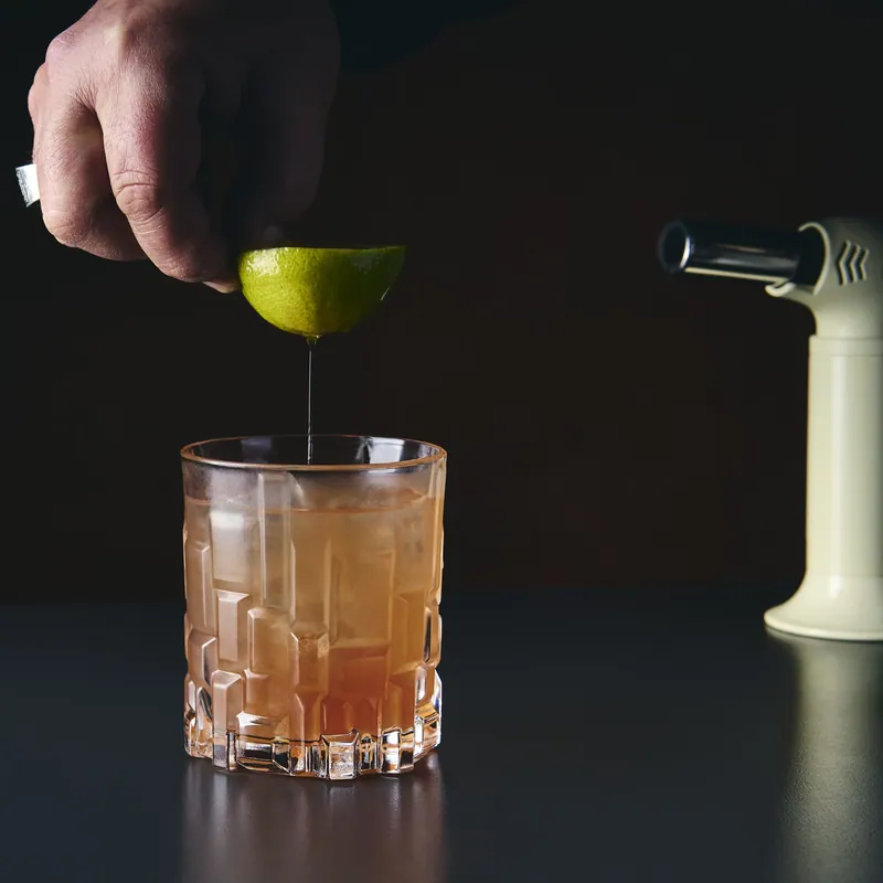 Mano che spreme un lime sopra un cocktail ambrato