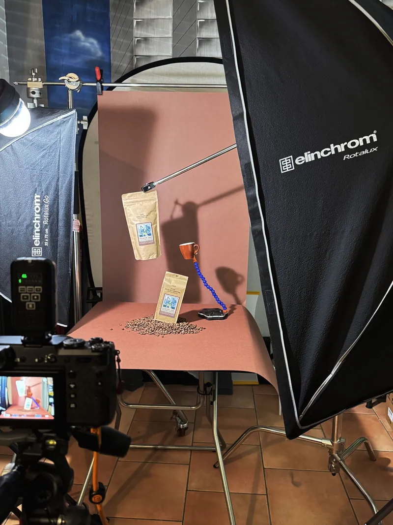Scena fotografica allestita con pacchetto di caffè, tazzina e chicchi, illuminata da softbox Elinchrom e supporti per oggetti sospesi su sfondo marrone.
